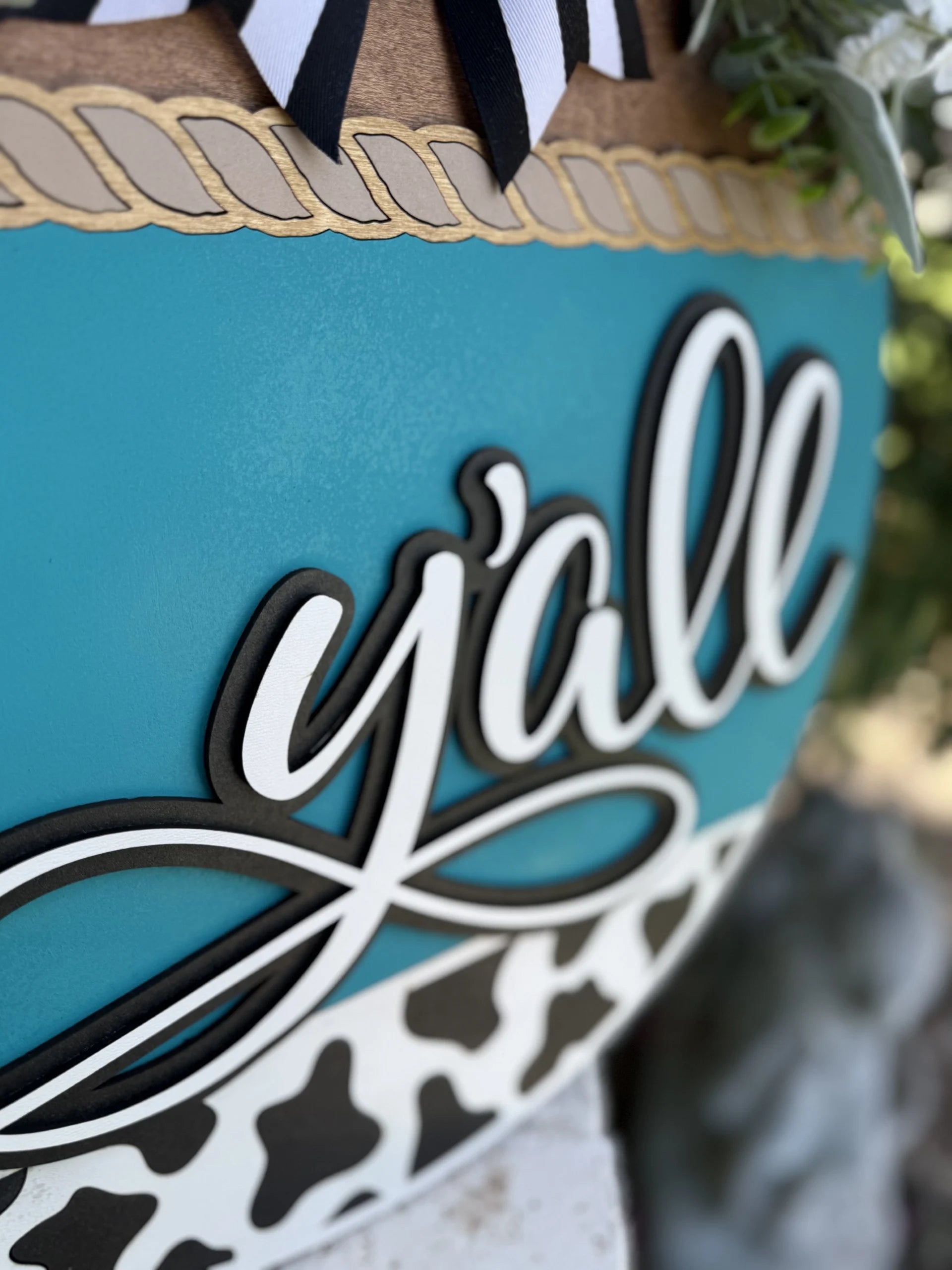 A close-up of a decorative sign featuring the word yall in bold, cursive letters on a blue background with rope detail and a black-and-white cow print pattern at the bottom.