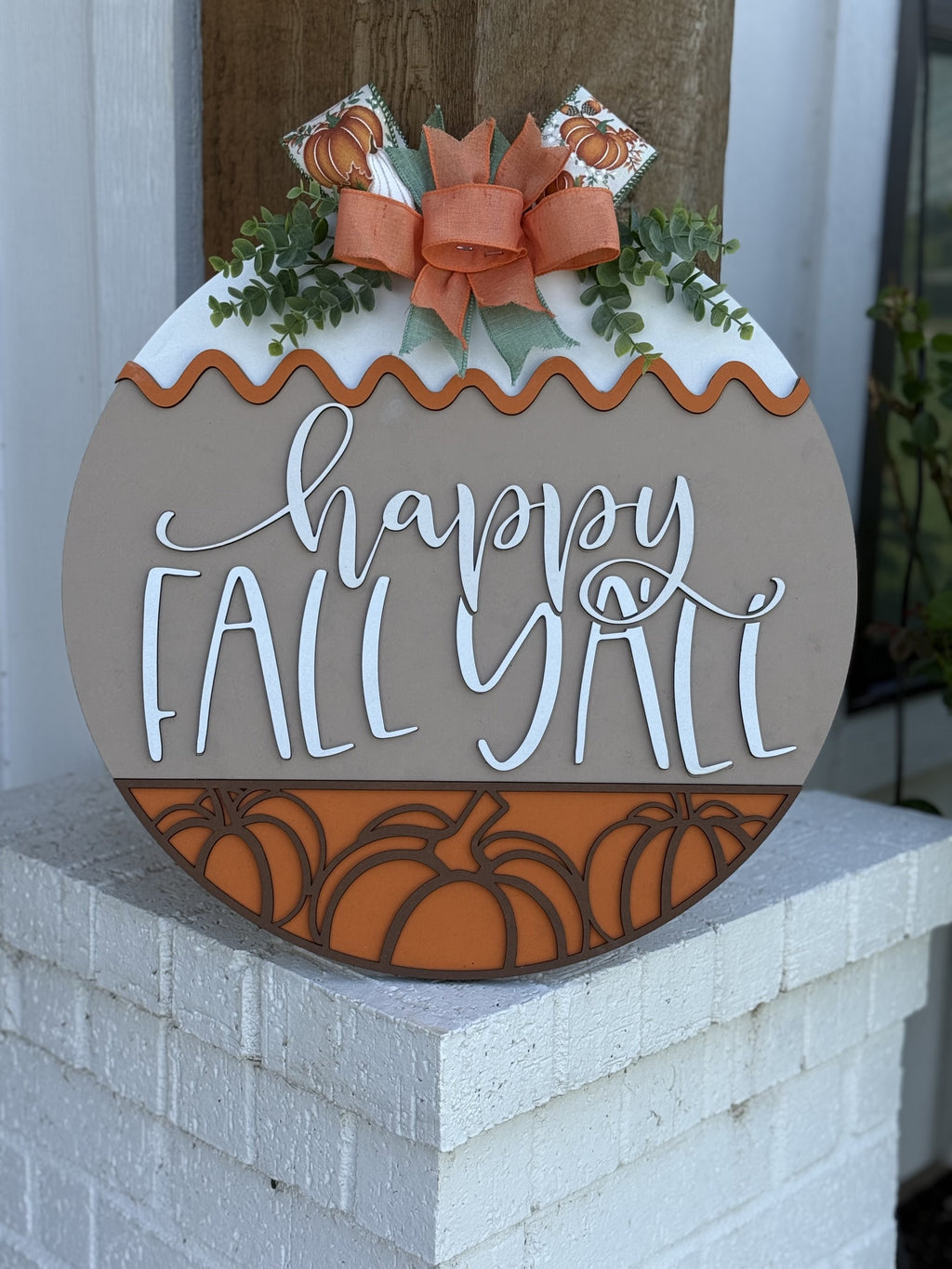 A round decorative sign with pumpkins, greenery, a bow, and the words happy FALL YALL in cursive, displayed on a white brick surface outdoors.