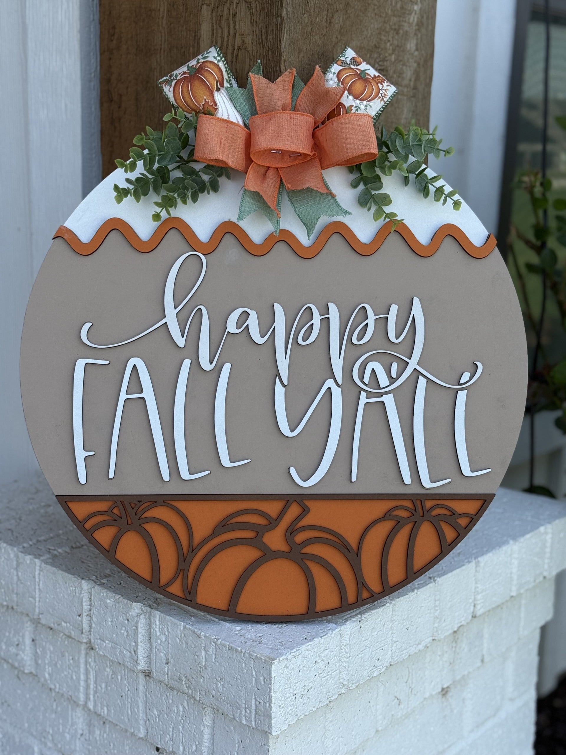 A round autumn-themed door sign with a bow, greenery, pumpkins, and the phrase “happy FALL Y’ALL” in decorative white script, leaning against a white brick surface.