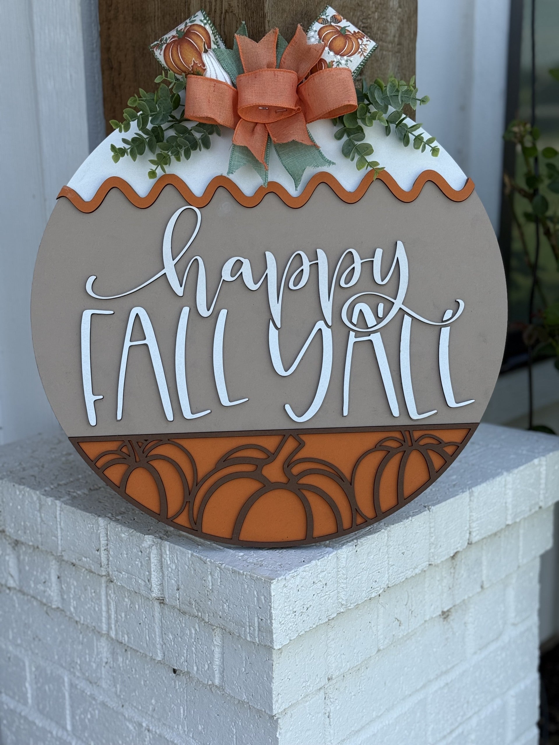A round wooden sign decorated with leaves, a bow, and pumpkins reads happy FALL YALL in cursive letters. The sign has orange and beige colors and is displayed outdoors on a white brick ledge.