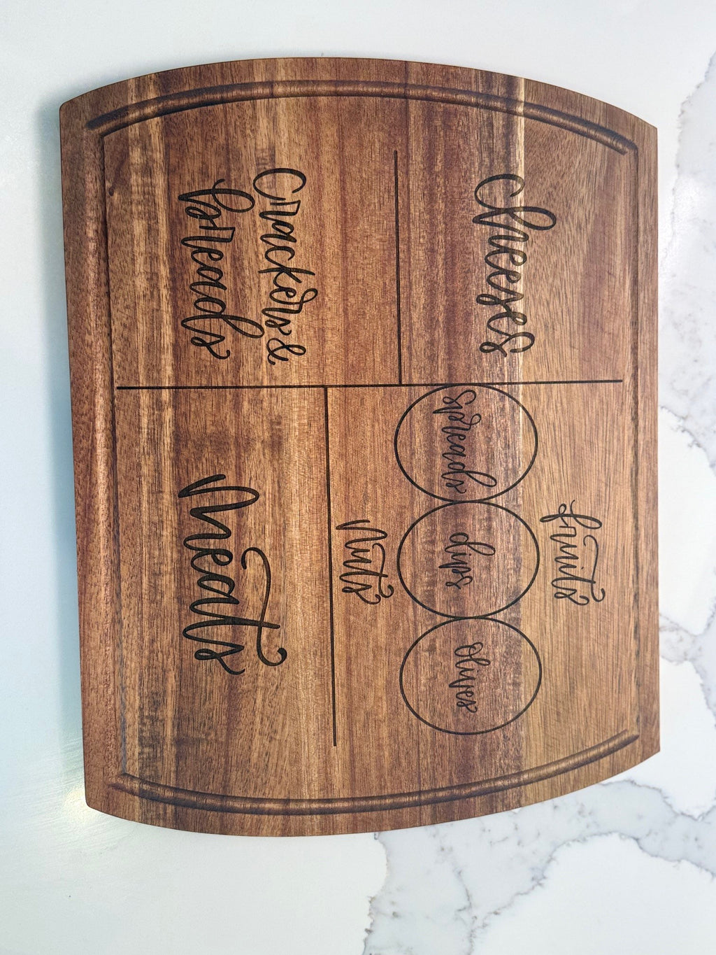 A wooden charcuterie board with engraved sections labeled cheeses, crackers & breads, meats, and circles for spreads, dips, and olives, placed on a white marble counter.