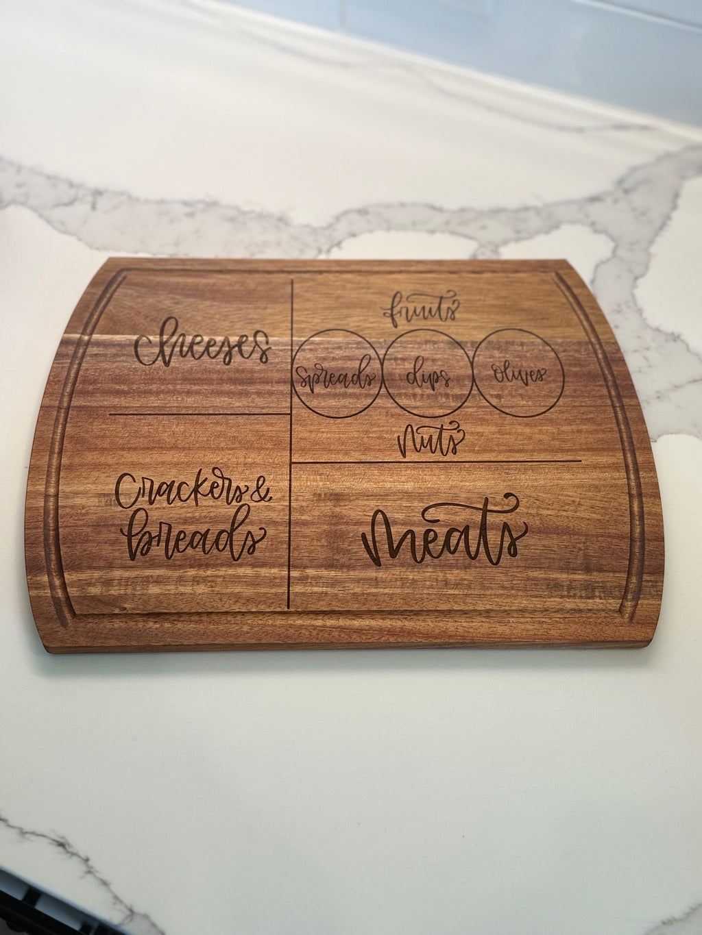 A wooden charcuterie board with engraved sections labeled cheeses, crackers & breads, meats, and circles for spreads, dips, and olives, placed on a white marble counter.