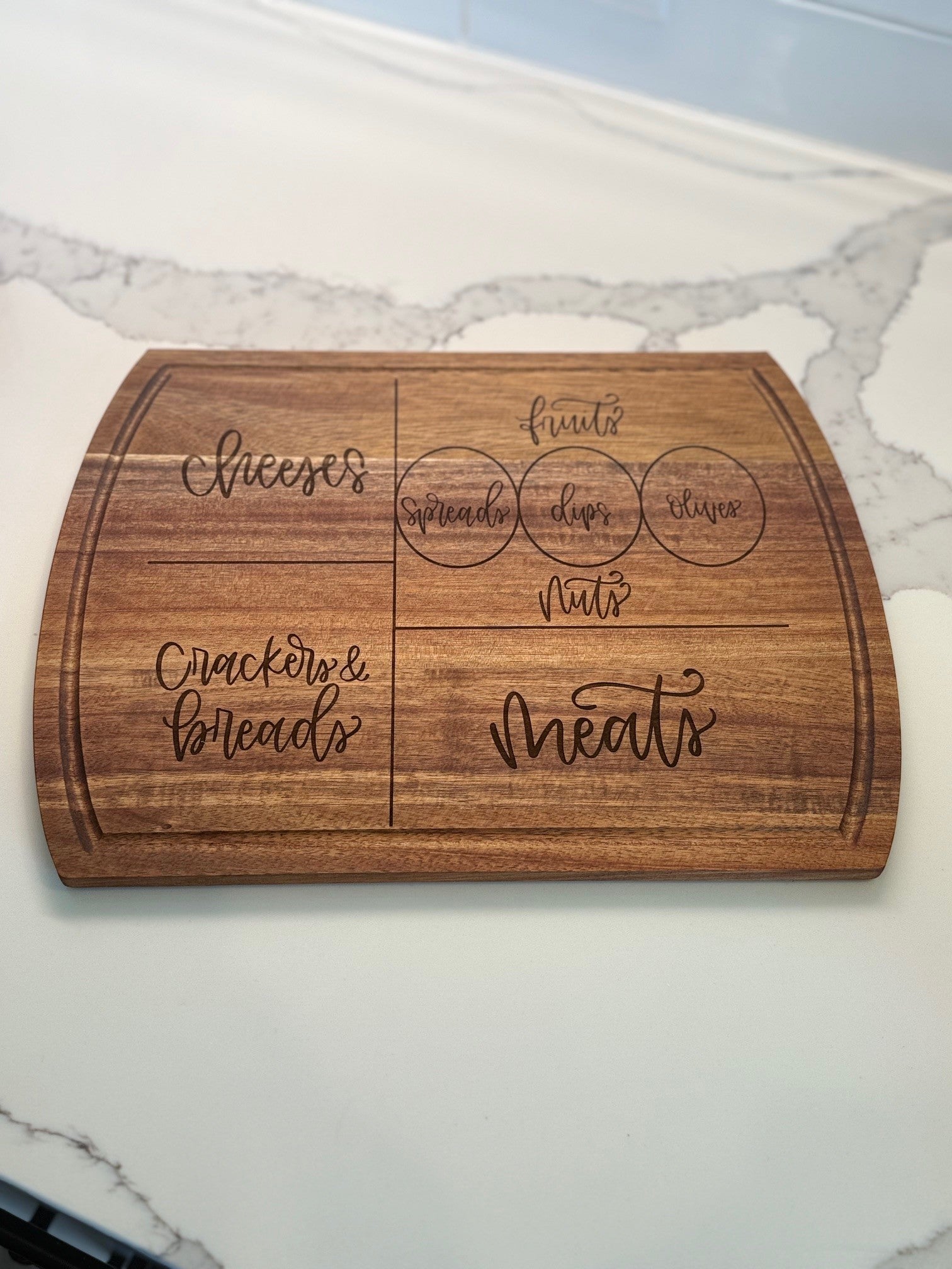 A wooden charcuterie board with engraved sections labeled cheeses, crackers & breads, meats, and circles for spreads, dips, and olives, placed on a white marble counter.