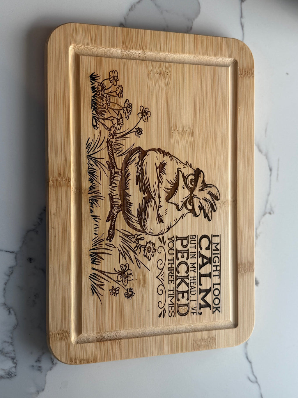 A wooden cutting board engraved with an illustration of a chicken sitting among flowers and grass, with the text: I might look calm, but in my head, Ive pecked you three times.