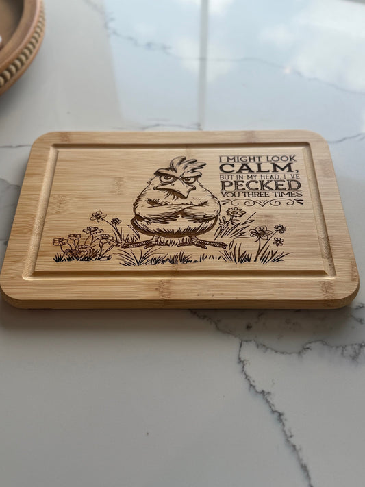 A wooden cutting board engraved with a cartoon chicken and the text, I might look calm, but in my head Ive pecked you three times, sitting on a marble countertop.