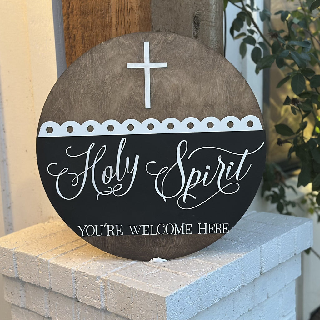 A circular wooden sign on a white brick ledge displays a cross at the top and the text Holy Spirit in elegant script, with YOURE WELCOME HERE in capital letters below. The sign has decorative cutouts and is set against a backdrop of a wooden pillar and leafy plant.
