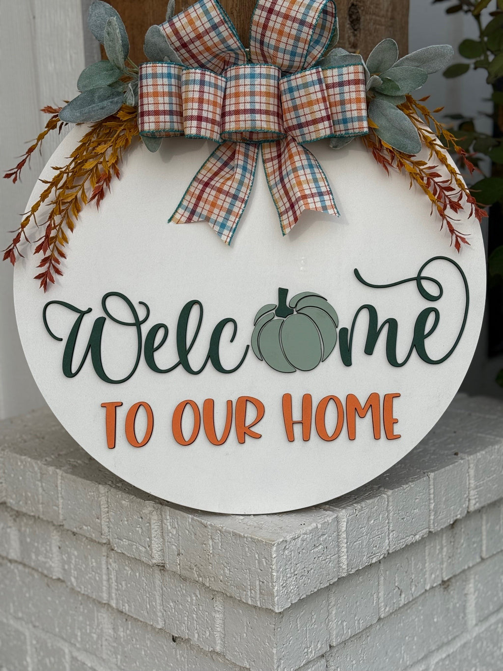 Round white sign with a plaid bow, greenery, and orange sprigs. It reads Welcome to our home, with a green pumpkin in place of the “o” in Welcome. The sign rests on a white brick ledge.
