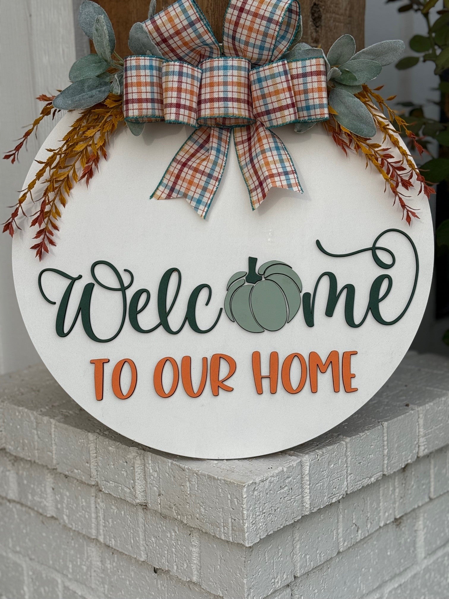 Round white sign with a plaid bow, greenery, and orange sprigs. It reads Welcome to our home, with a green pumpkin in place of the “o” in Welcome. The sign rests on a white brick ledge.