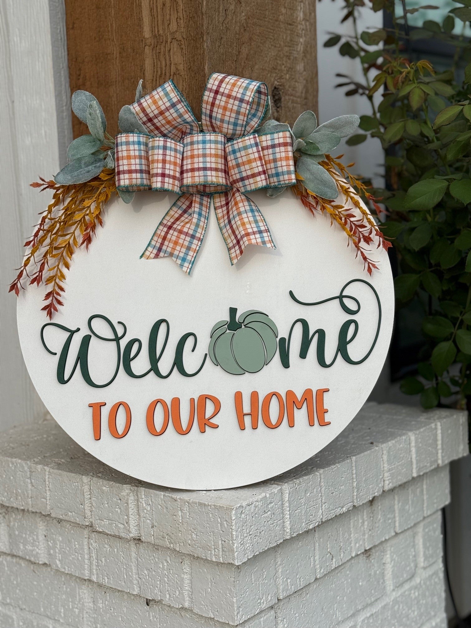 Round white sign with a plaid bow, greenery, and orange sprigs. It reads Welcome to our home, with a green pumpkin in place of the “o” in Welcome. The sign rests on a white brick ledge.