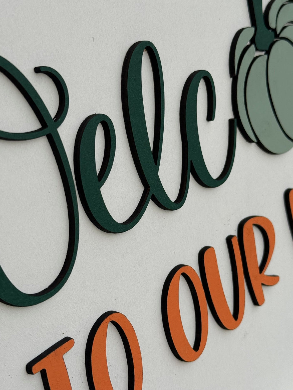 Round white sign with a plaid bow, greenery, and orange sprigs. It reads Welcome to our home, with a green pumpkin in place of the “o” in Welcome. The sign rests on a white brick ledge.