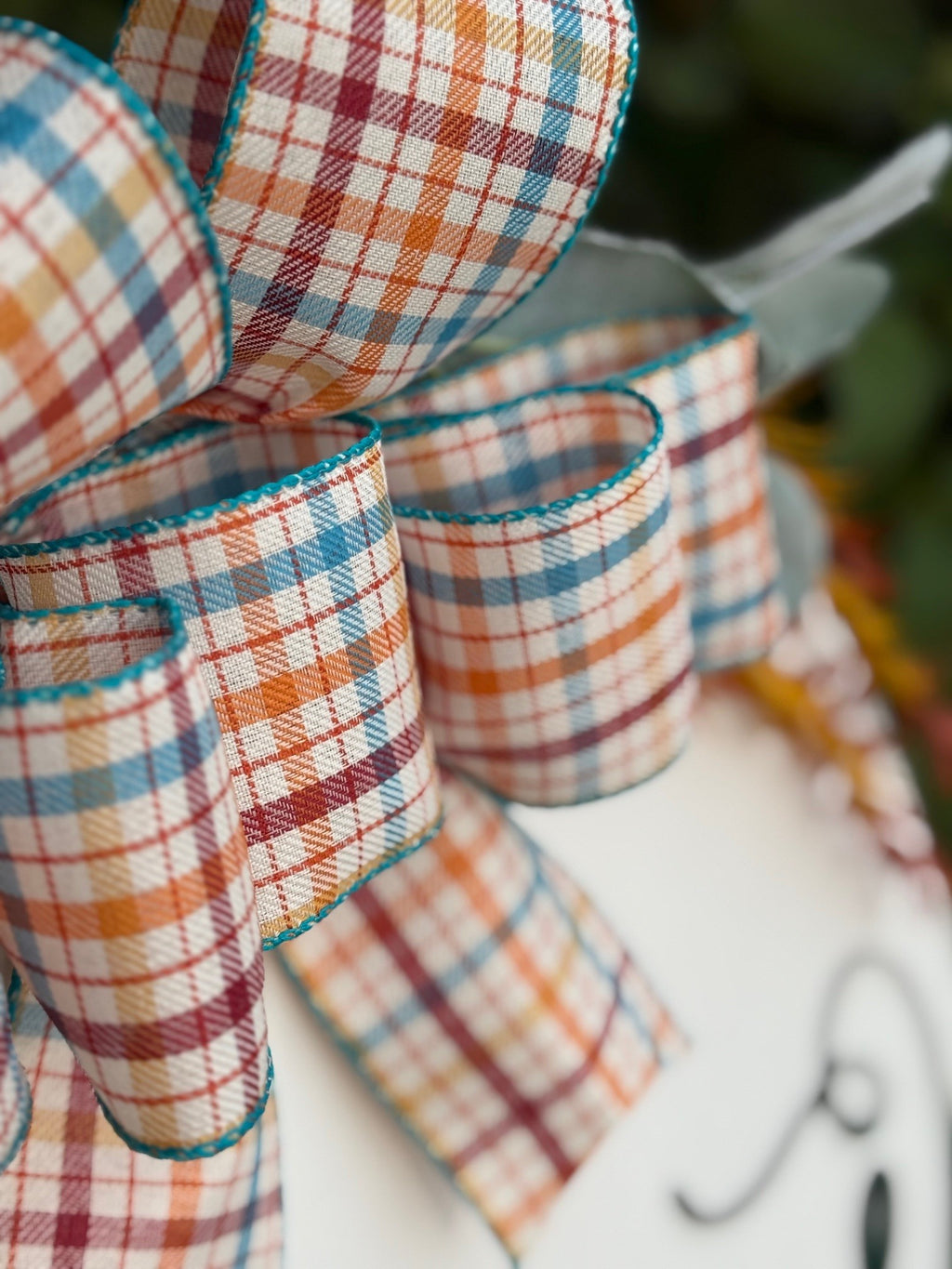 Round white sign with a plaid bow, greenery, and orange sprigs. It reads Welcome to our home, with a green pumpkin in place of the “o” in Welcome. The sign rests on a white brick ledge.