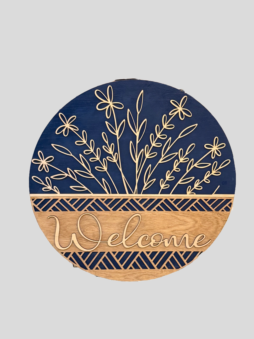 The Floral Basket Welcome Sign is a rustic wooden door hanger. It features the word Welcome in cursive on a wooden background, topped with a blue section showcasing an engraved floral basket design. Geometric patterns elegantly separate the sections.