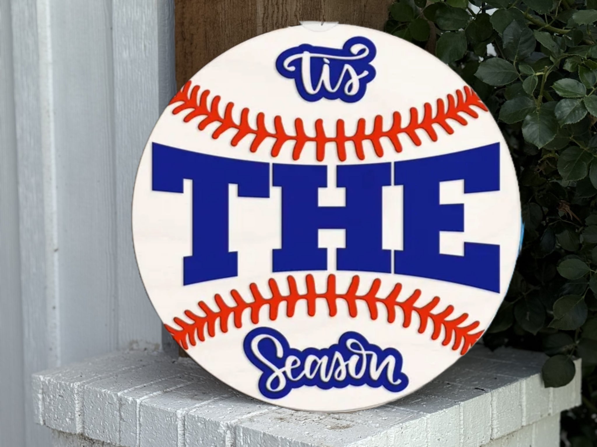A round sign resembling a baseball with red stitching reads “Tis THE Season” in blue and white lettering. The sign is propped against a white brick surface next to plants and a wooden post.