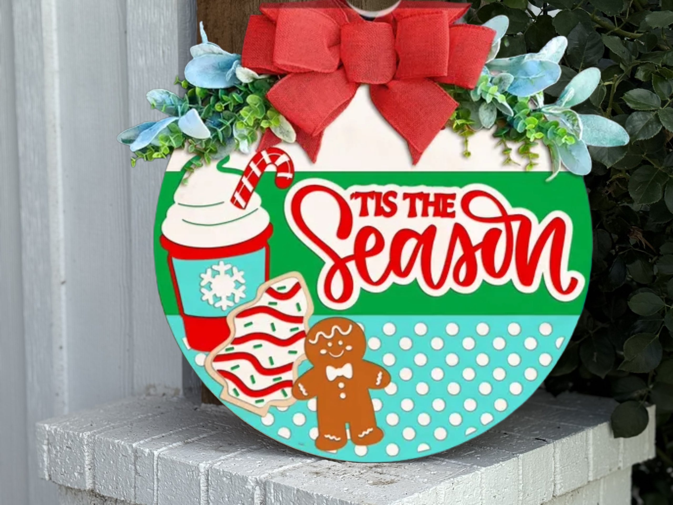 A festive sign shaped like an ornament features a red bow, greenery, a coffee cup with a candy cane, decorated cookies, a gingerbread figure, and the text “’Tis the Season” in red script.