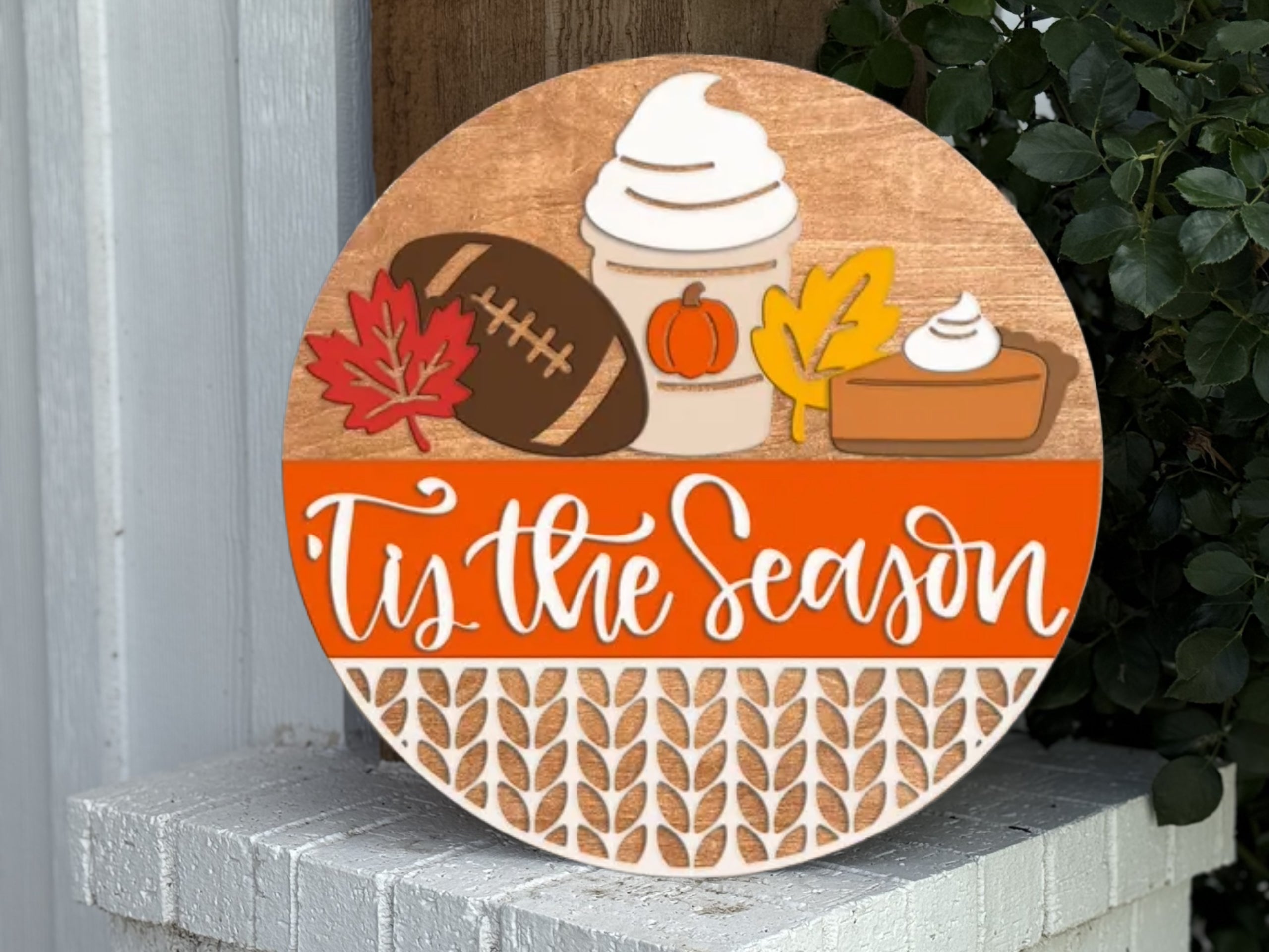 Round wooden sign decorated with a football, red and yellow leaves, a pumpkin spice drink, and a slice of pie above the words “’Tis the Season” in white script, placed outdoors near green foliage.