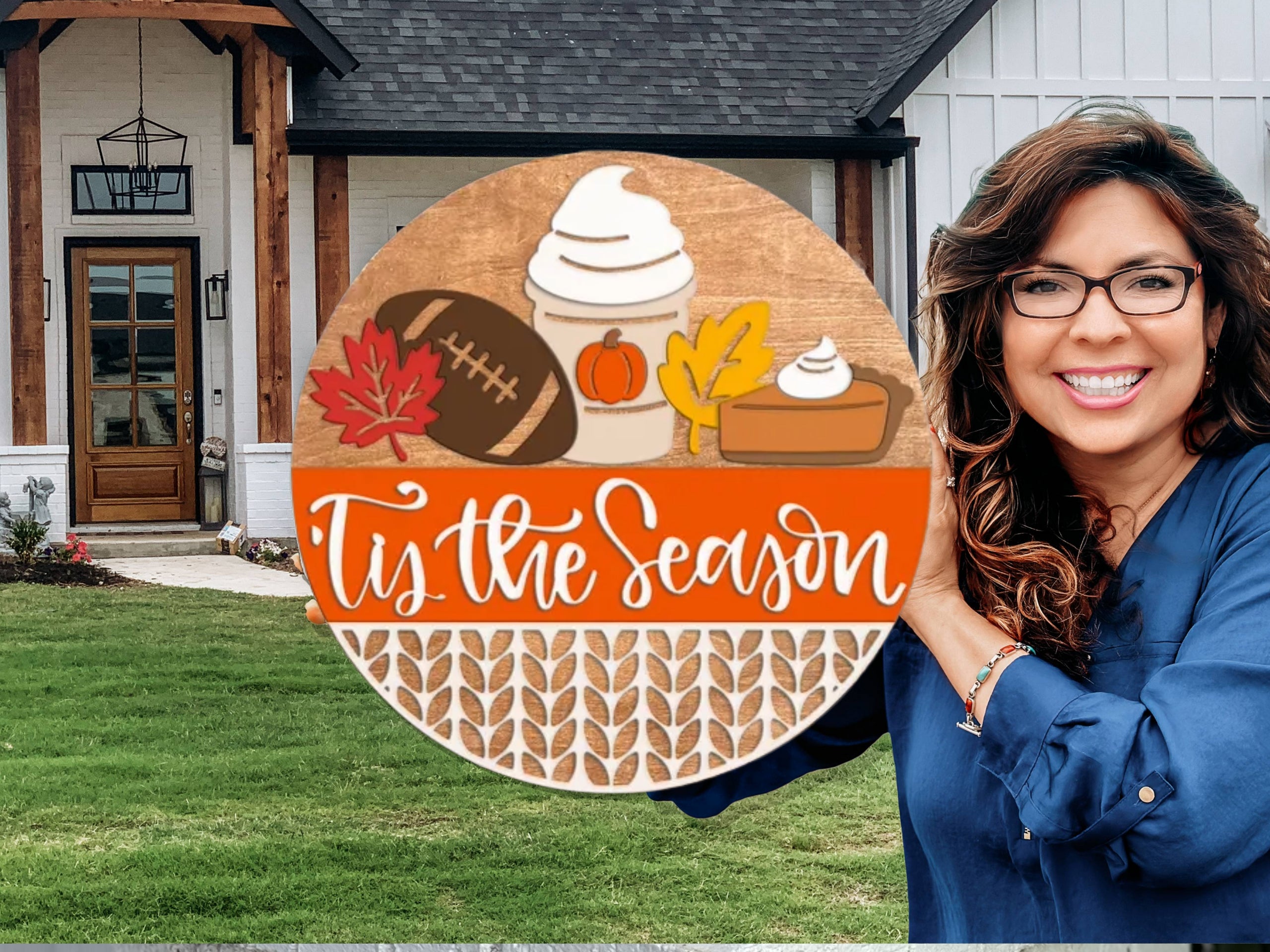 A smiling woman with long brown hair and glasses holds a round wooden sign decorated with fall items, reading Tis the Season, standing in front of a modern house with a lawn.