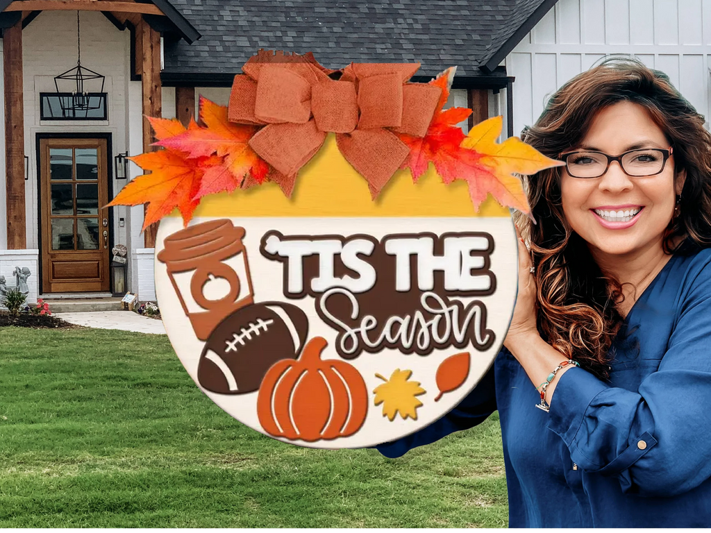 A smiling woman in glasses holds a decorative round sign with a bow, autumn leaves, a coffee cup, football, pumpkin, and leaves. The sign reads TIS THE Season. A modern house with a lawn is in the background.
