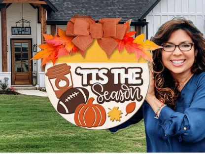 A smiling woman in glasses holds a decorative round sign with a bow, autumn leaves, a coffee cup, football, pumpkin, and leaves. The sign reads TIS THE Season. A modern house with a lawn is in the background.