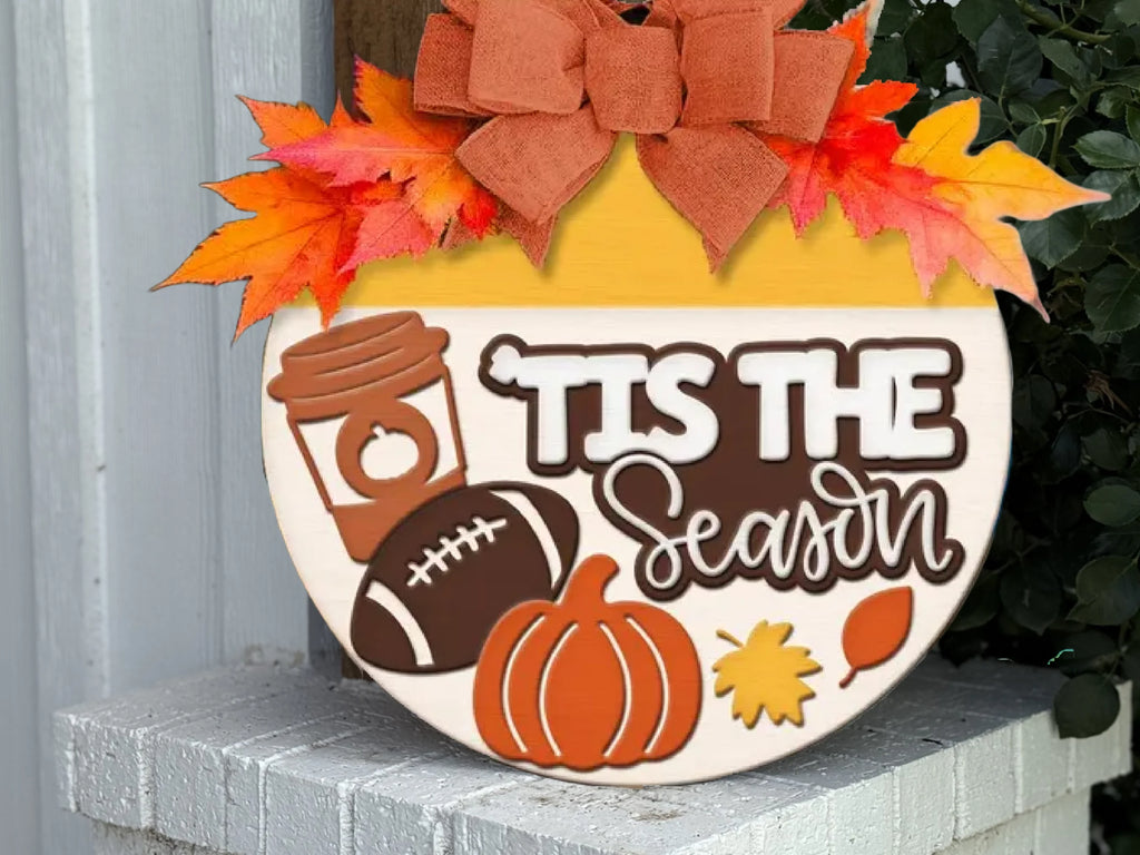 A round fall-themed sign on a white brick ledge features a bow, autumn leaves, a coffee cup, football, pumpkin, and leaf, with the phrase Tis the Season in bold and script letters.