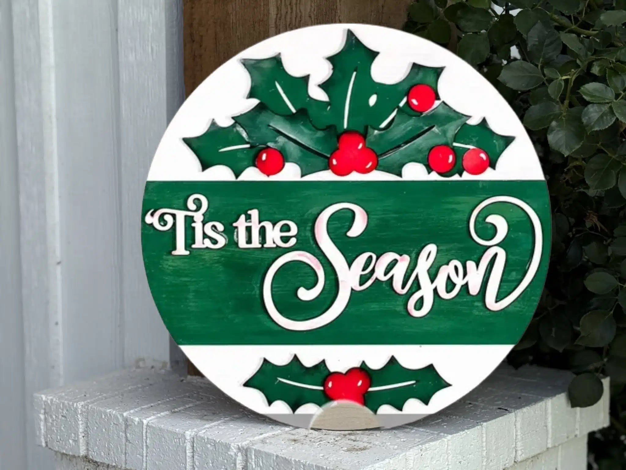 A round sign with green holly leaves and red berries reads Tis the Season in white script. The sign is displayed on a white brick ledge next to a leafy plant and a white wall.