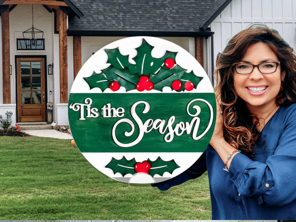 A smiling woman with wavy hair and glasses holds a round sign that reads Tis the Season with holly decorations, standing in front of a modern house with wood and white accents.