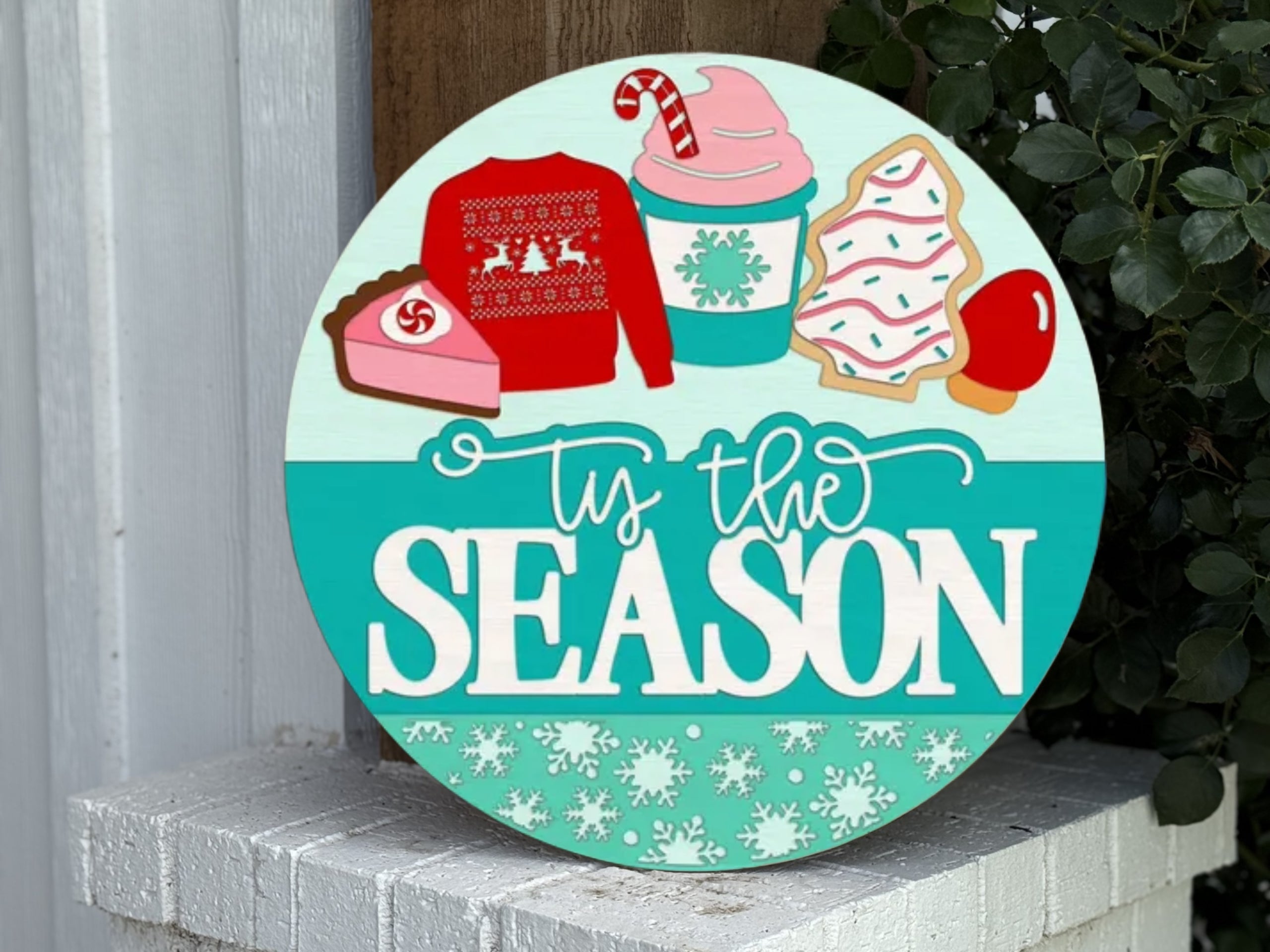 A round sign reading “’Tis the Season” with festive images above, including a holiday sweater, pink pie slice, mint drink with a snowflake, decorated cookies, and a caramel apple. Snowflakes border the bottom of the sign.