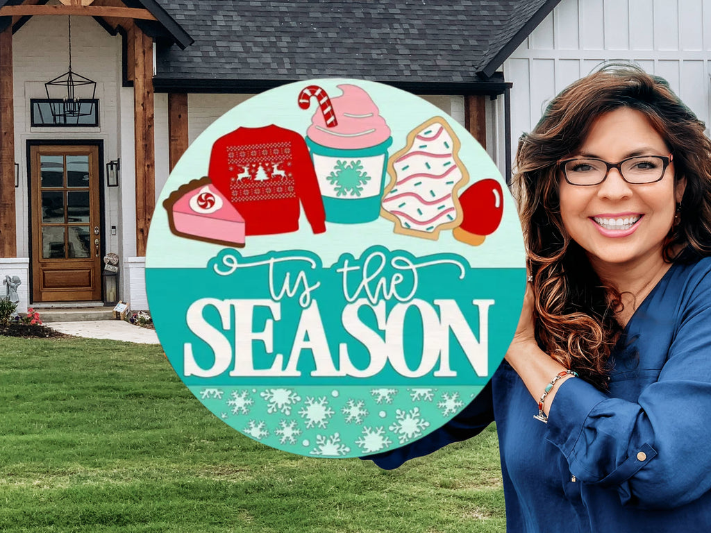 A smiling woman with long brown hair and glasses holds a round sign in front of a house. The sign features holiday treats, a sweater, and the phrase ’tis the season in festive colors.