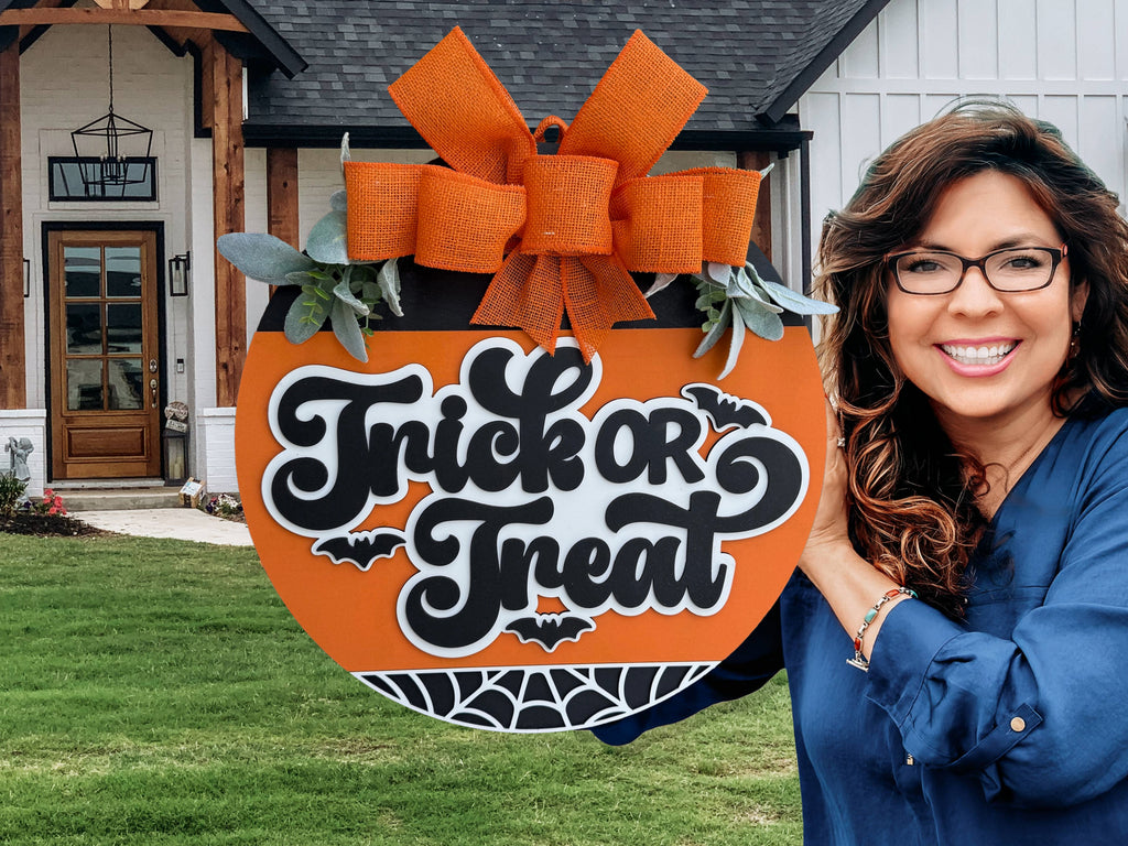 A smiling woman with wavy brown hair holds a large, round Trick or Treat Halloween sign with an orange bow and black-and-white lettering, standing outside in front of a modern house.
