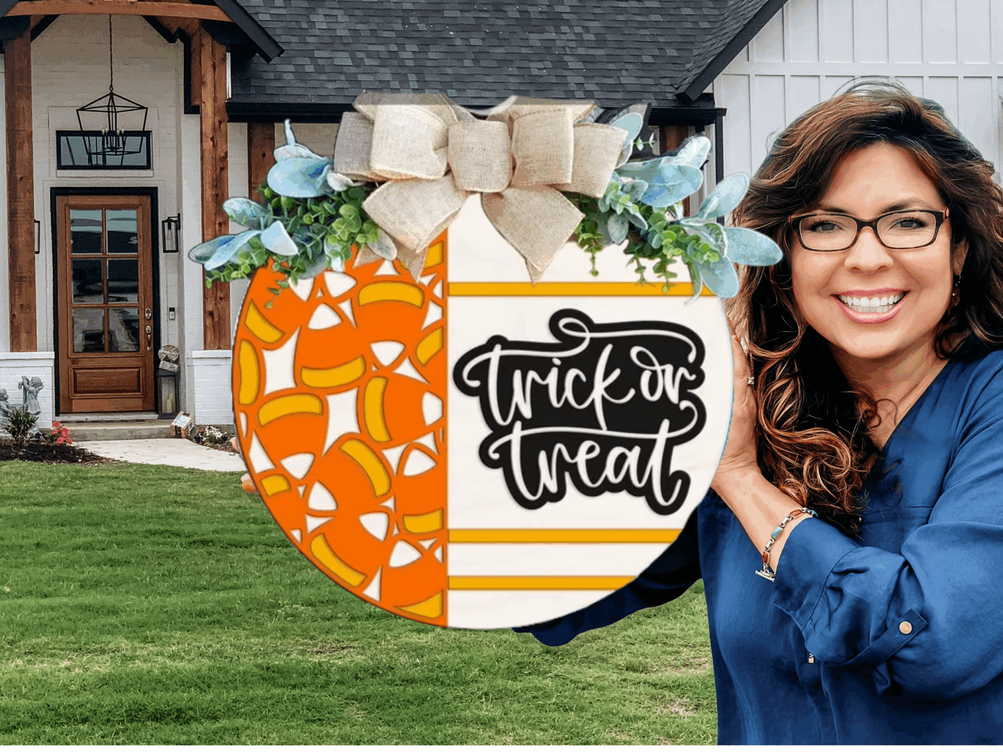 A smiling woman with glasses holds a round Halloween sign decorated with candy corn patterns, greenery, a burlap bow, and the words trick or treat in front of a modern house with a lawn.