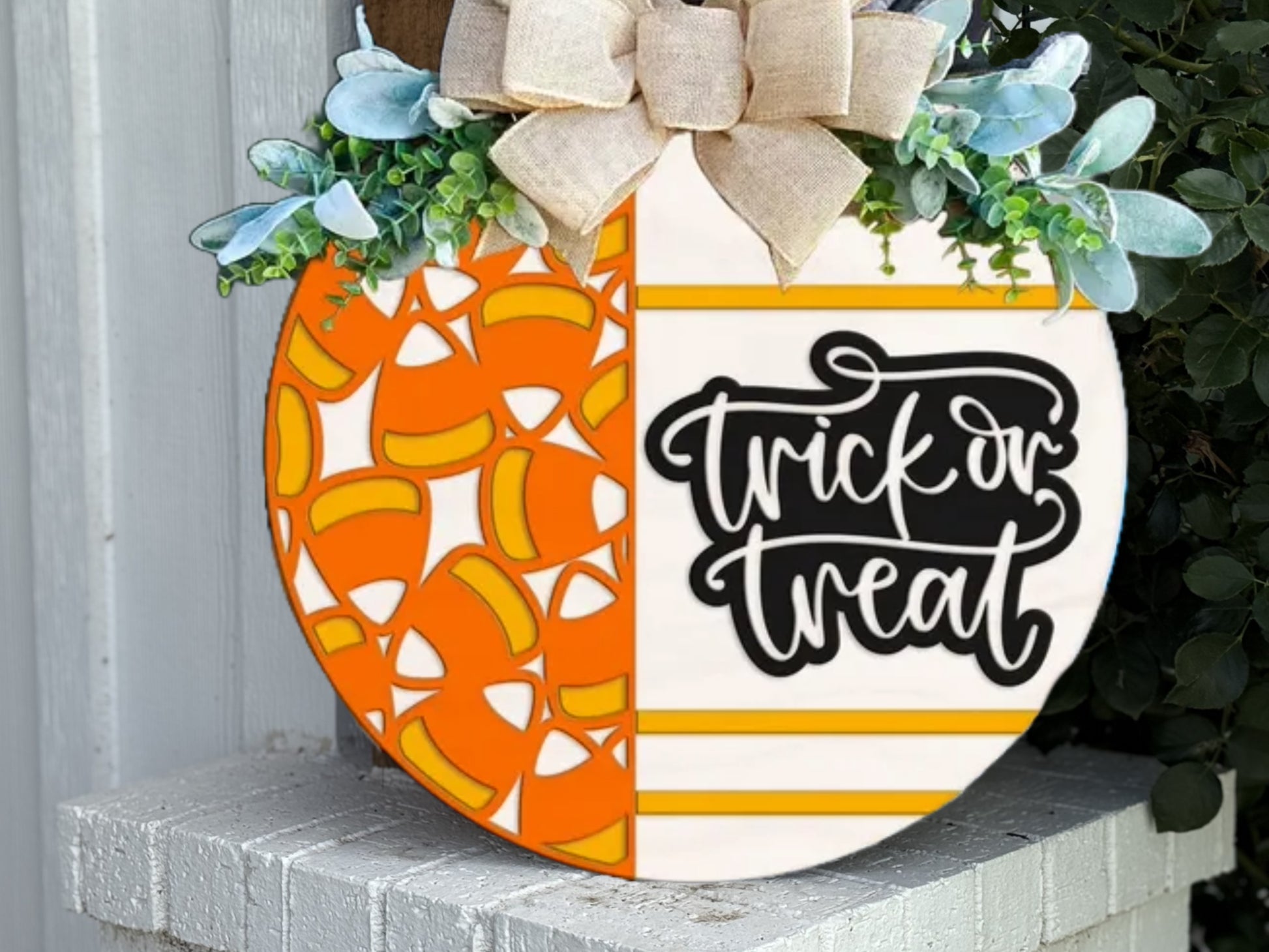 A round Halloween sign decorated with candy corn designs on one half and “trick or treat” text on the other, topped with a bow and greenery, leaning against a white wall.