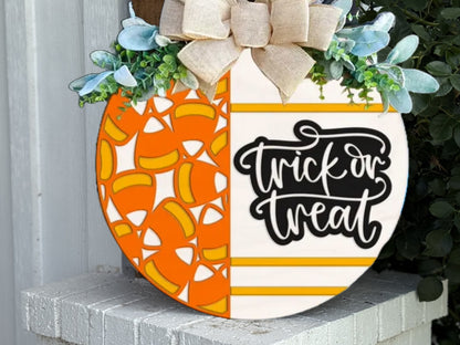 A round Halloween sign decorated with candy corn designs on one half and “trick or treat” text on the other, topped with a bow and greenery, leaning against a white wall.
