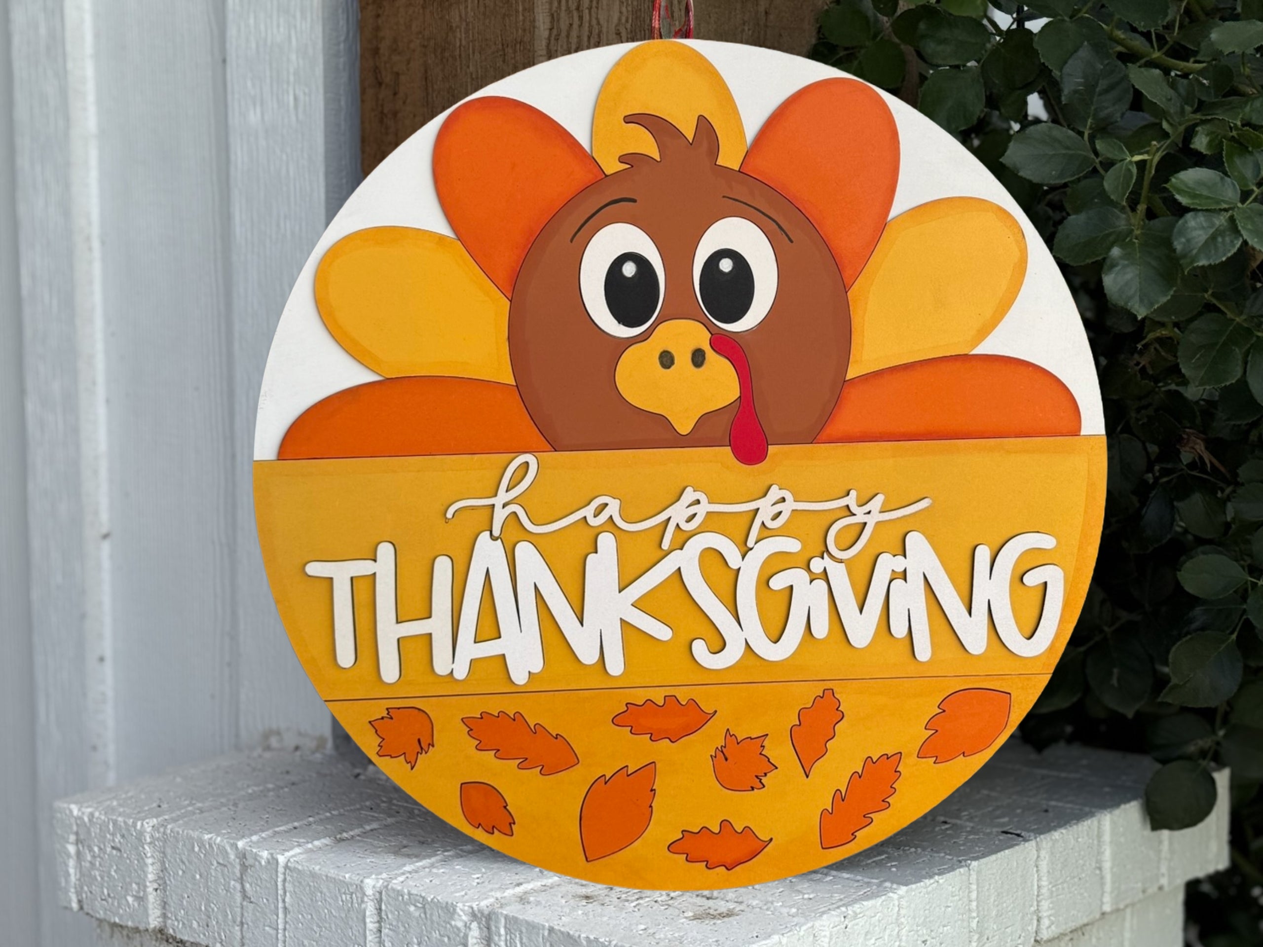 A round Thanksgiving sign featuring a cartoon turkey with a red wattle and yellow beak, colorful feathers, and the words Happy Thanksgiving surrounded by orange leaves.