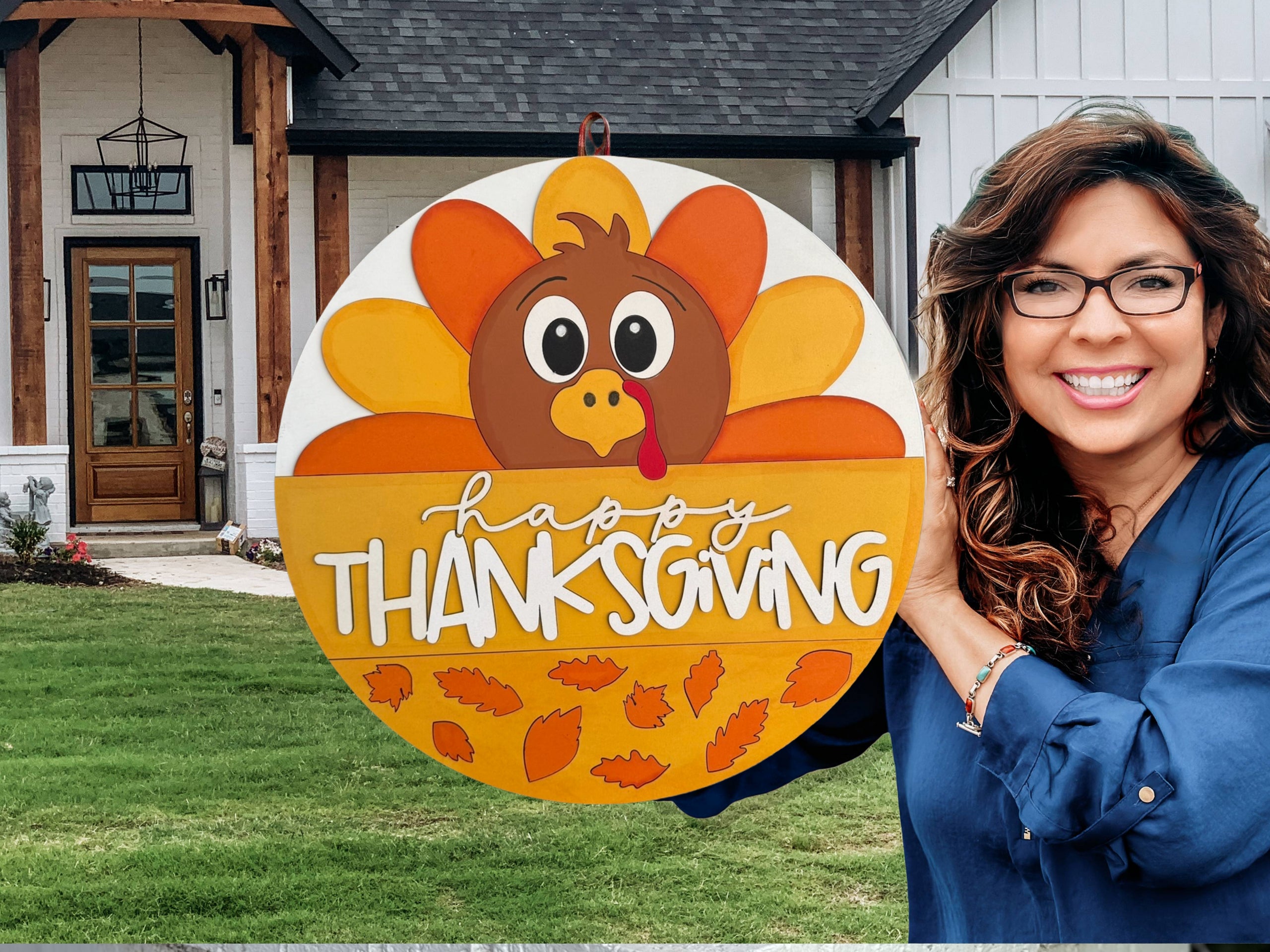 A smiling woman with long, wavy brown hair and glasses holds a round Happy Thanksgiving sign featuring a cartoon turkey and autumn leaves, standing outside a modern house with a green lawn.