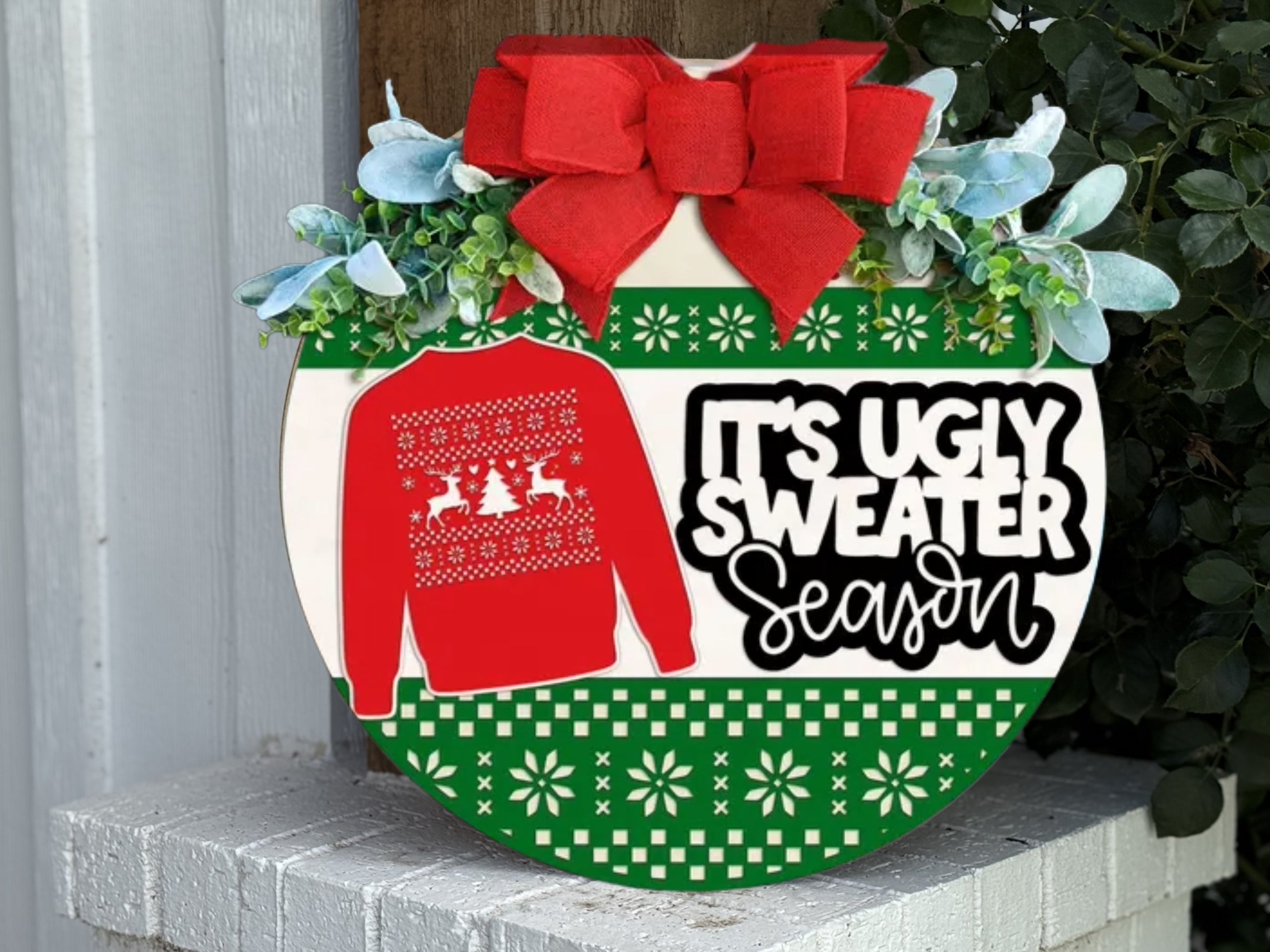 A festive sign with a red bow, greenery, and a red sweater graphic reads Its Ugly Sweater Season on a green and white patterned background, displayed on a white brick ledge.