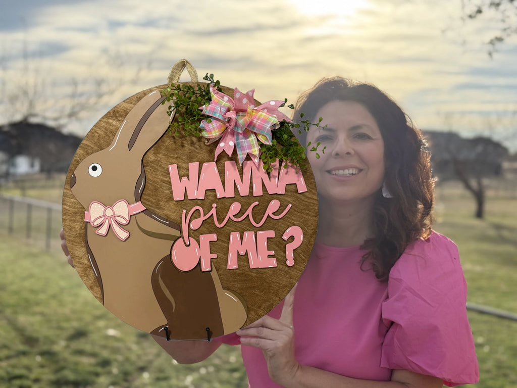 A woman in a pink shirt holds a circular wooden Easter decoration with a brown rabbit design. The text on the sign reads Wanna piece of me? in pink letters. A small bow and greenery adorn the top. The background shows a grassy park with trees and a sunny sky.
