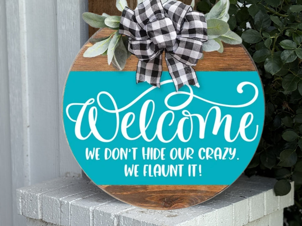 The Welcome to Our Home Wooden Door Hanger features a black-and-white plaid bow, green leaves, and the message: Welcome. We don’t hide our crazy. We flaunt it! This rustic round farmhouse sign is perfect porch decor.