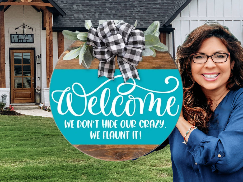 A smiling woman holds a round teal sign with a black-and-white plaid bow, displaying the text: Welcome. We dont hide our crazy. We flaunt it! She stands in front of a modern house with a wooden door.