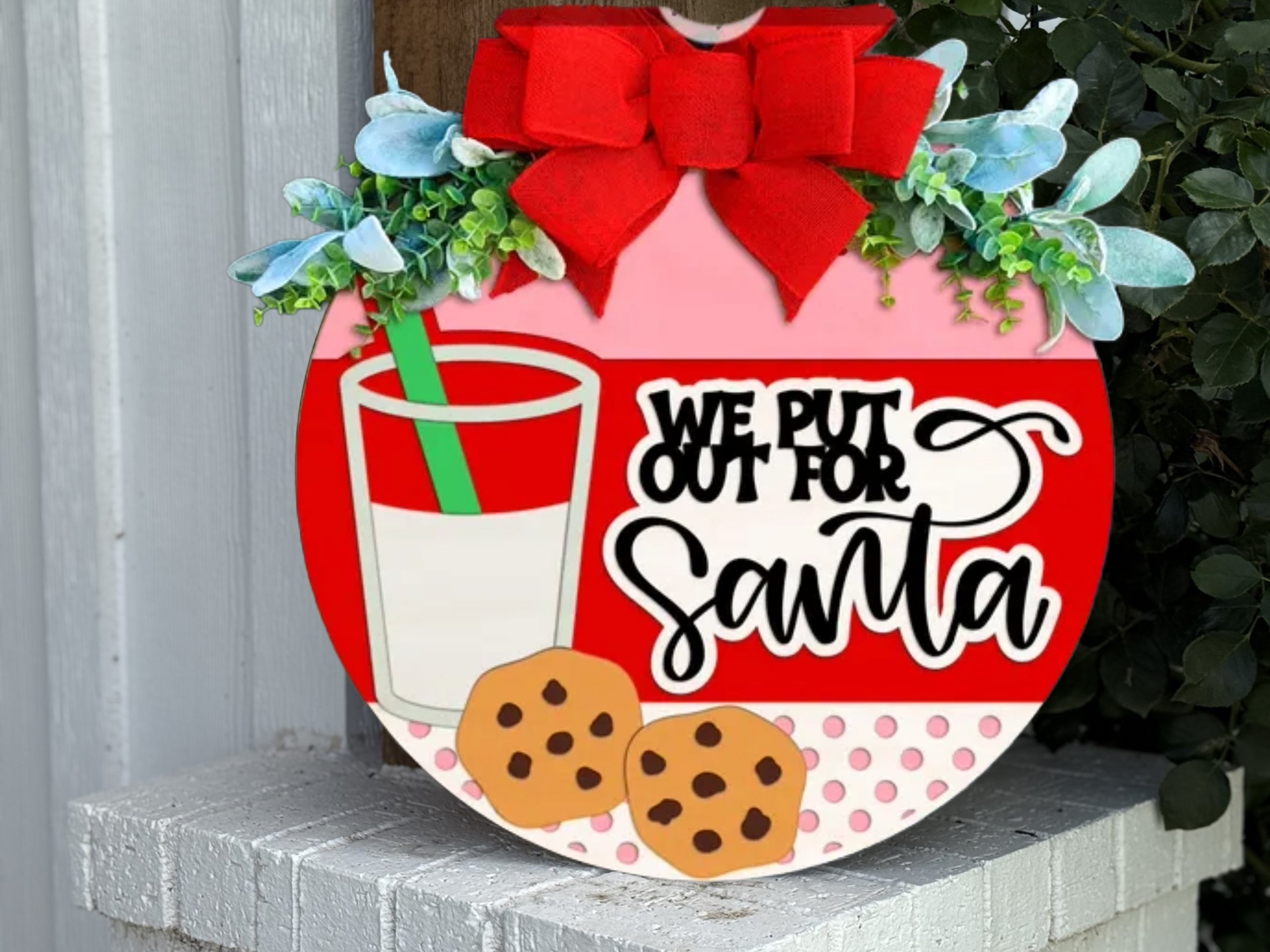A festive round sign decorated with a red bow, greenery, a glass of milk, two cookies, and the text “We put out for Santa” in playful fonts, set against a white brick background.