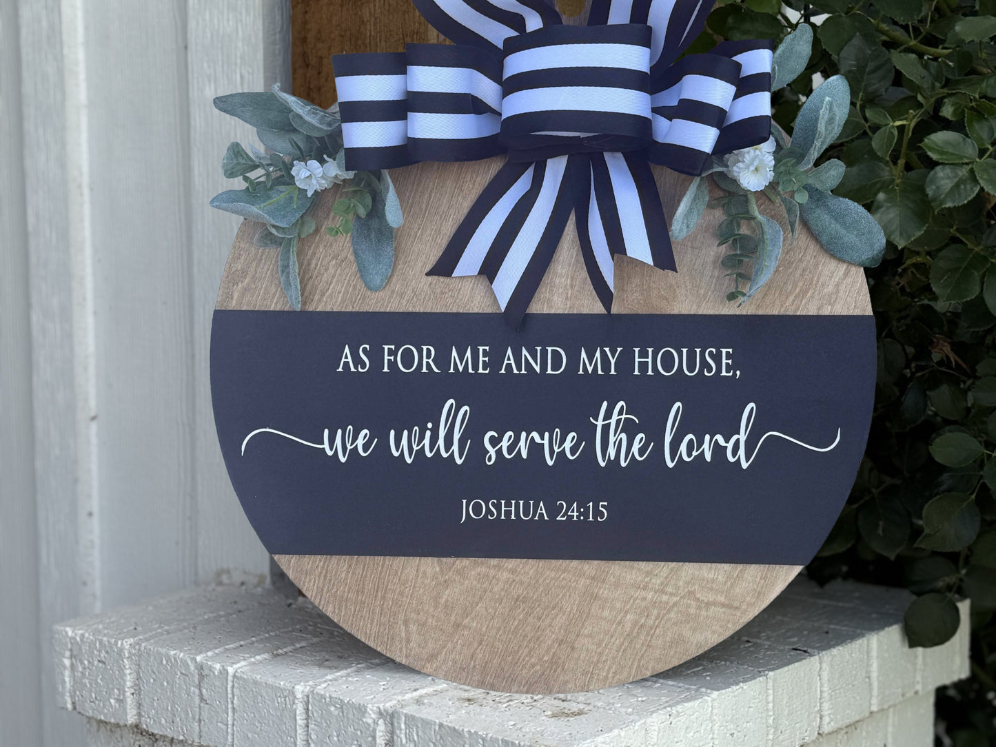 A round wooden sign with blue and white ribbon and greenery, displaying the Bible verse: As for me and my house, we will serve the Lord. Joshua 24:15, leaning on a white brick ledge.