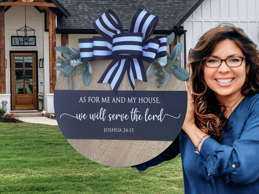 A smiling woman stands outside a house, holding a round sign decorated with a blue-and-white bow and greenery. The sign reads, As for me and my house, we will serve the Lord. Joshua 24:15.