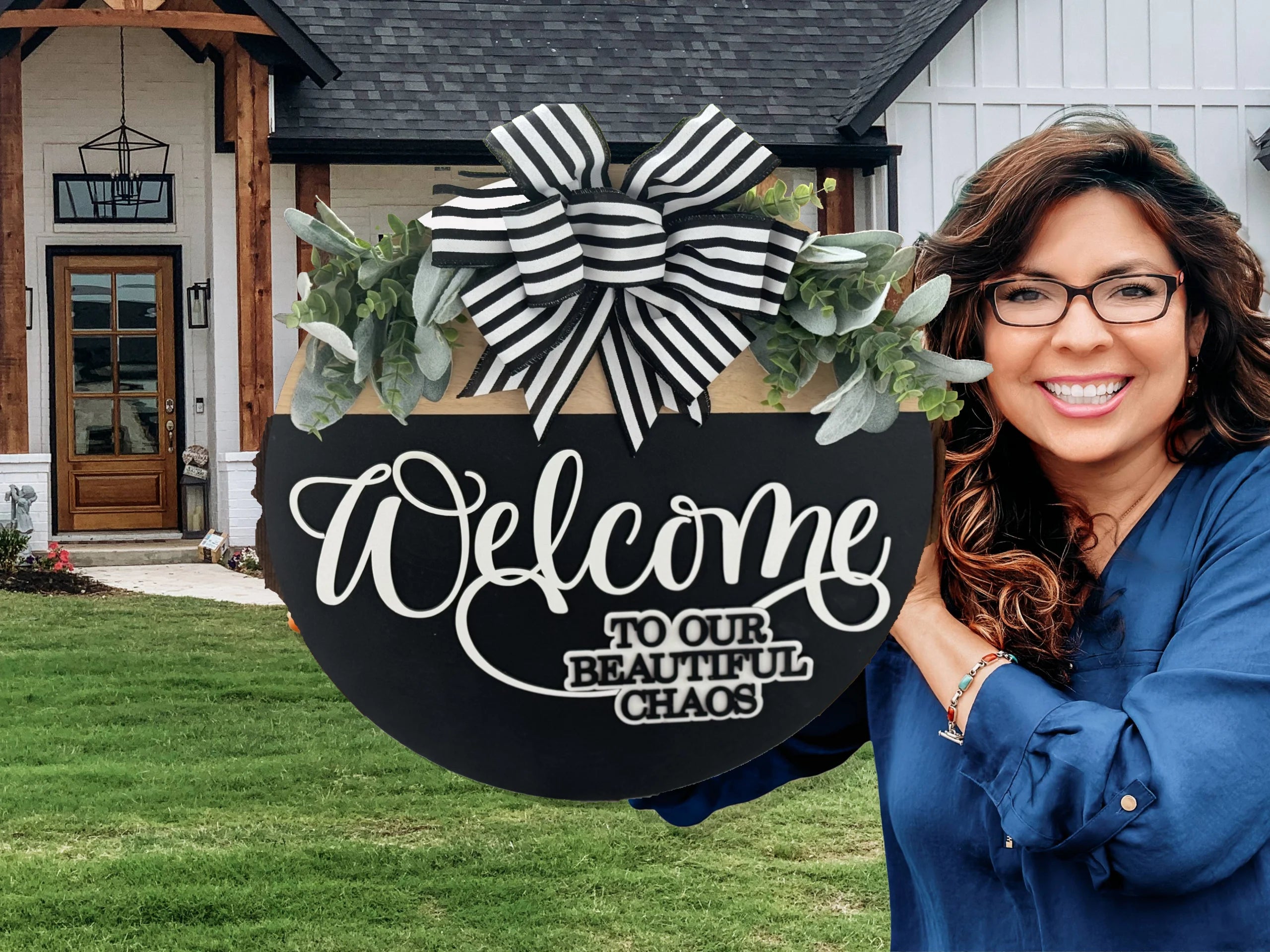 A person in a blue shirt is smiling and holding a decorative sign. The sign is black with white cursive text that reads, Welcome to our beautiful chaos. It features a black-and-white striped bow and green foliage. The background shows a grassy lawn and a modern house entrance.
