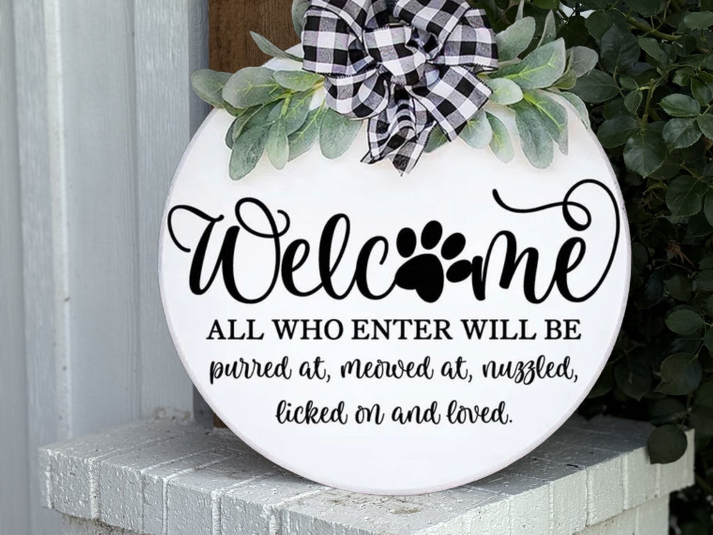 A round white sign with a black and white plaid bow and greenery reads: “Welcome. All who enter will be purred at, meowed at, nuzzled, licked on and loved.” The word Welcome has a paw print in place of the o.