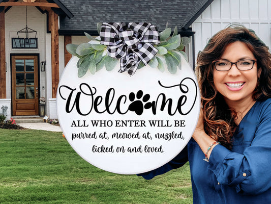A smiling woman holds a round sign with a bow and leaves, reading “Welcome. All who enter will be purred at, meowed at, nuzzled, licked on and loved.” She stands outside a modern farmhouse-style home.