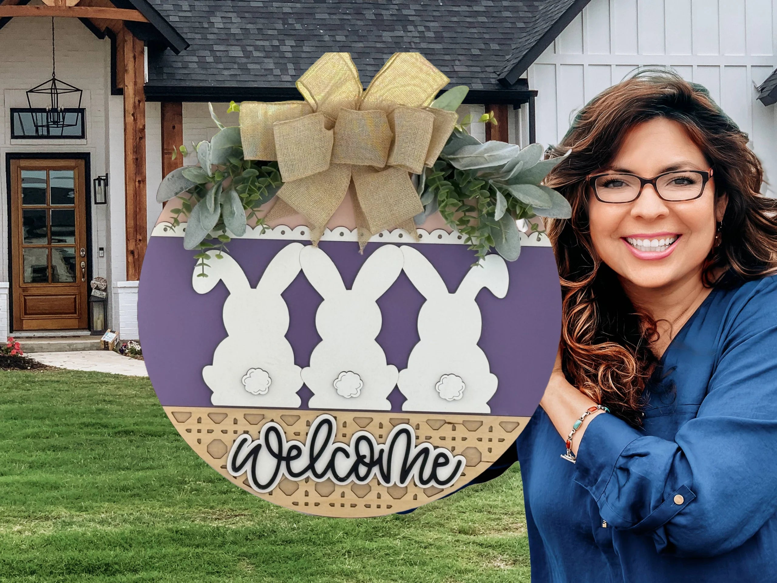 A smiling woman with wavy brown hair and glasses holds a large, round Easter-themed welcome sign decorated with three white bunny silhouettes, greenery, and a gold bow, standing outside a modern house with white siding and wood accents.