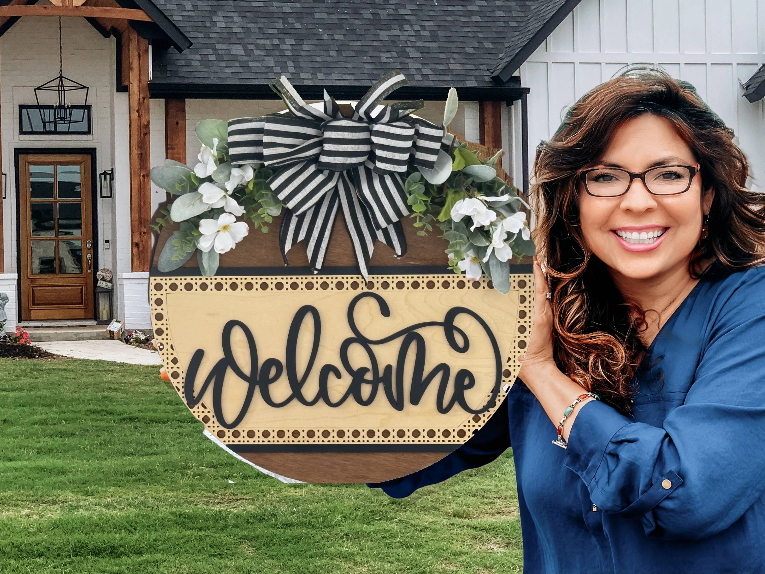 A smiling woman with long brown hair and glasses stands outside a modern house, holding a round wooden “Welcome” sign decorated with black-and-white striped ribbon, greenery, and white flowers. The background shows a green lawn and front porch.