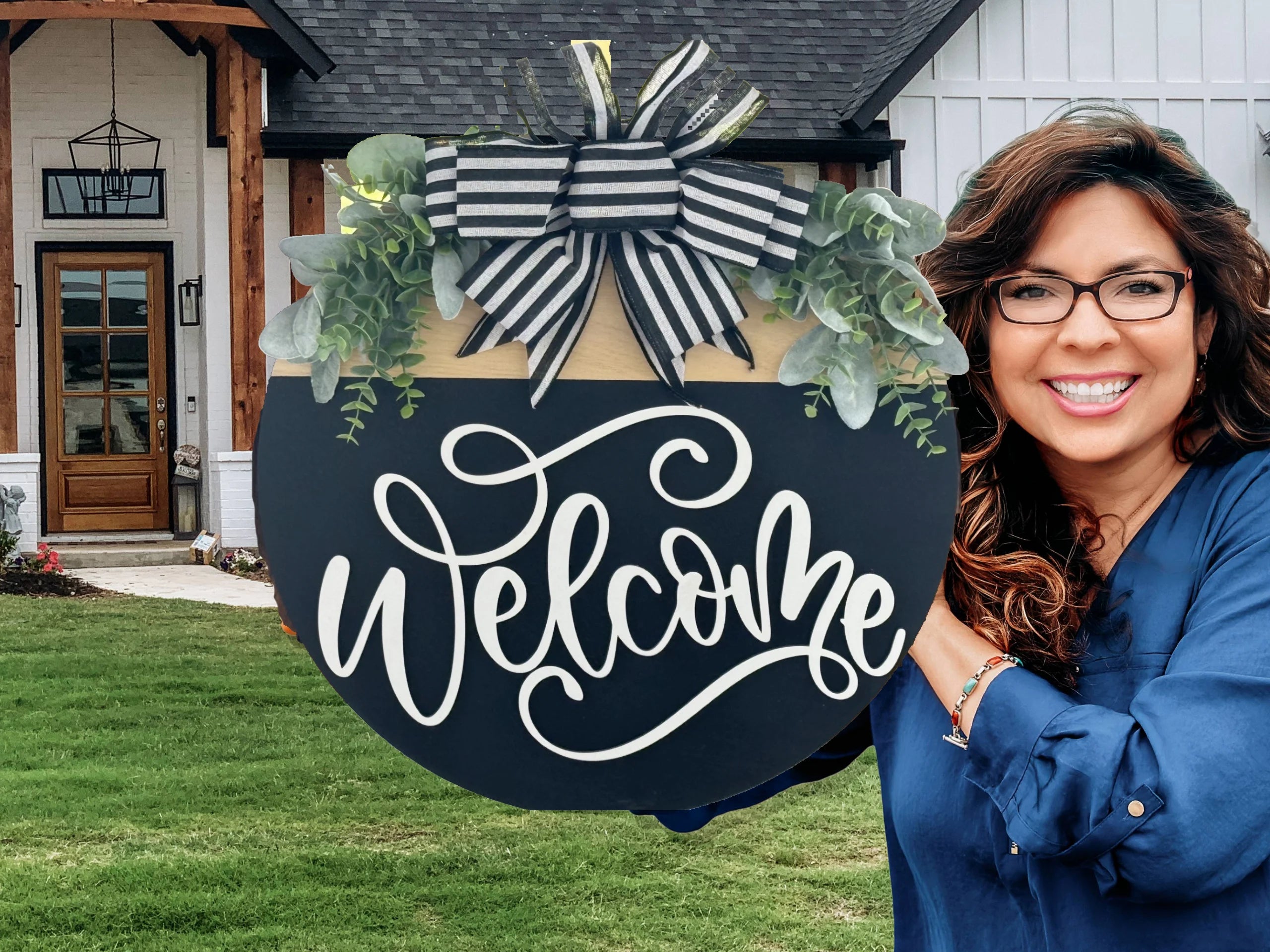 A smiling woman with long wavy hair and glasses stands outside a house, holding a large round Welcome sign decorated with striped ribbon, faux greenery, and elegant white script. The house has white siding, a wooden door, and a manicured lawn.