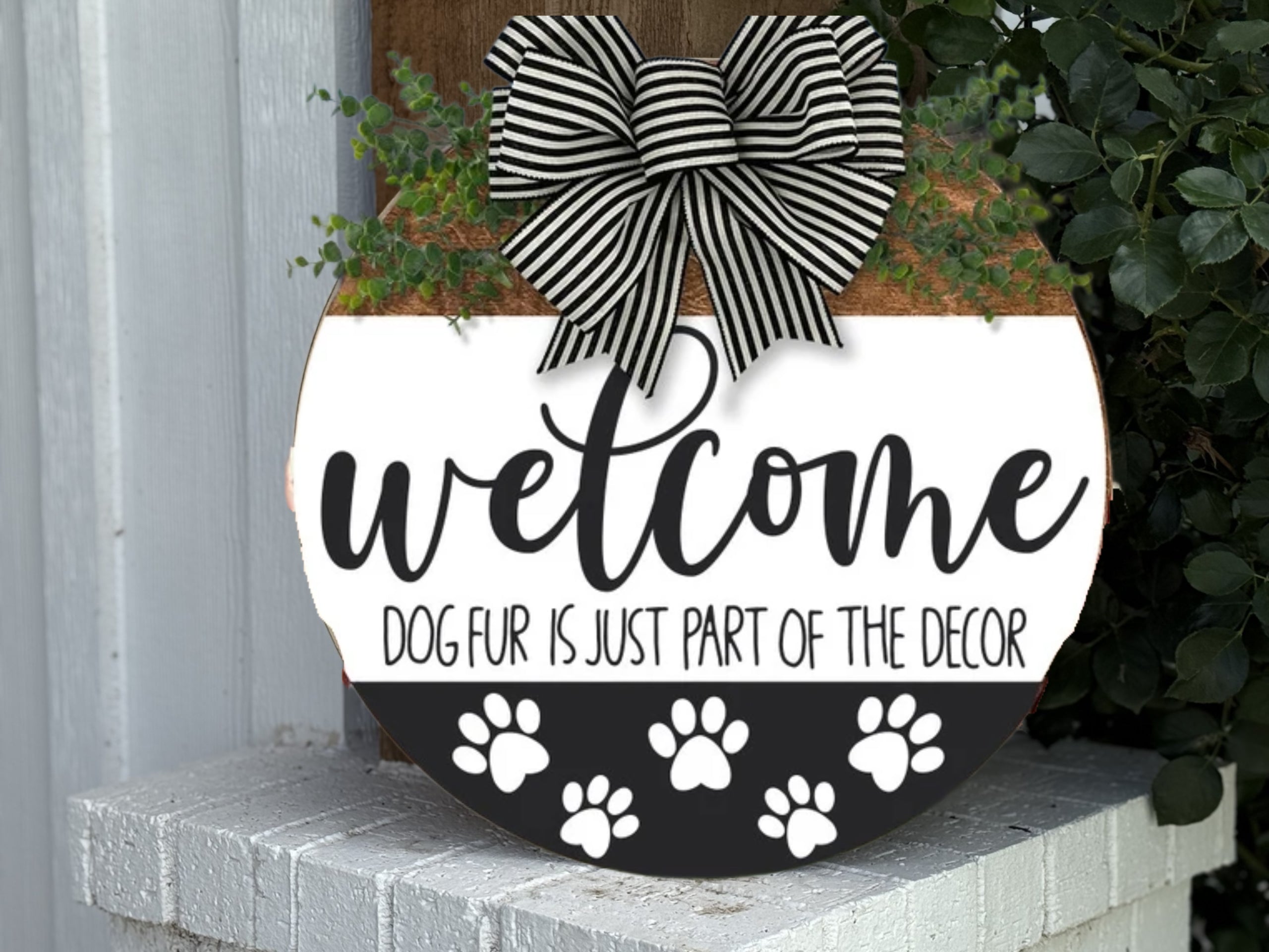 A round welcome sign with a black-and-white striped bow, decorated with paw prints and the text Welcome. Dog fur is just part of the decor, hanging on a white brick wall with green plants nearby.