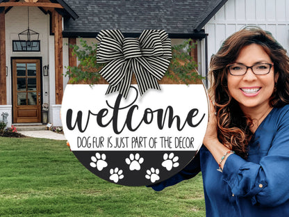 A smiling woman holding a round sign with a black-and-white striped bow reads, Welcome. Dog fur is just part of the decor, with paw prints. She stands outside a modern house with a lawn and wooden door.