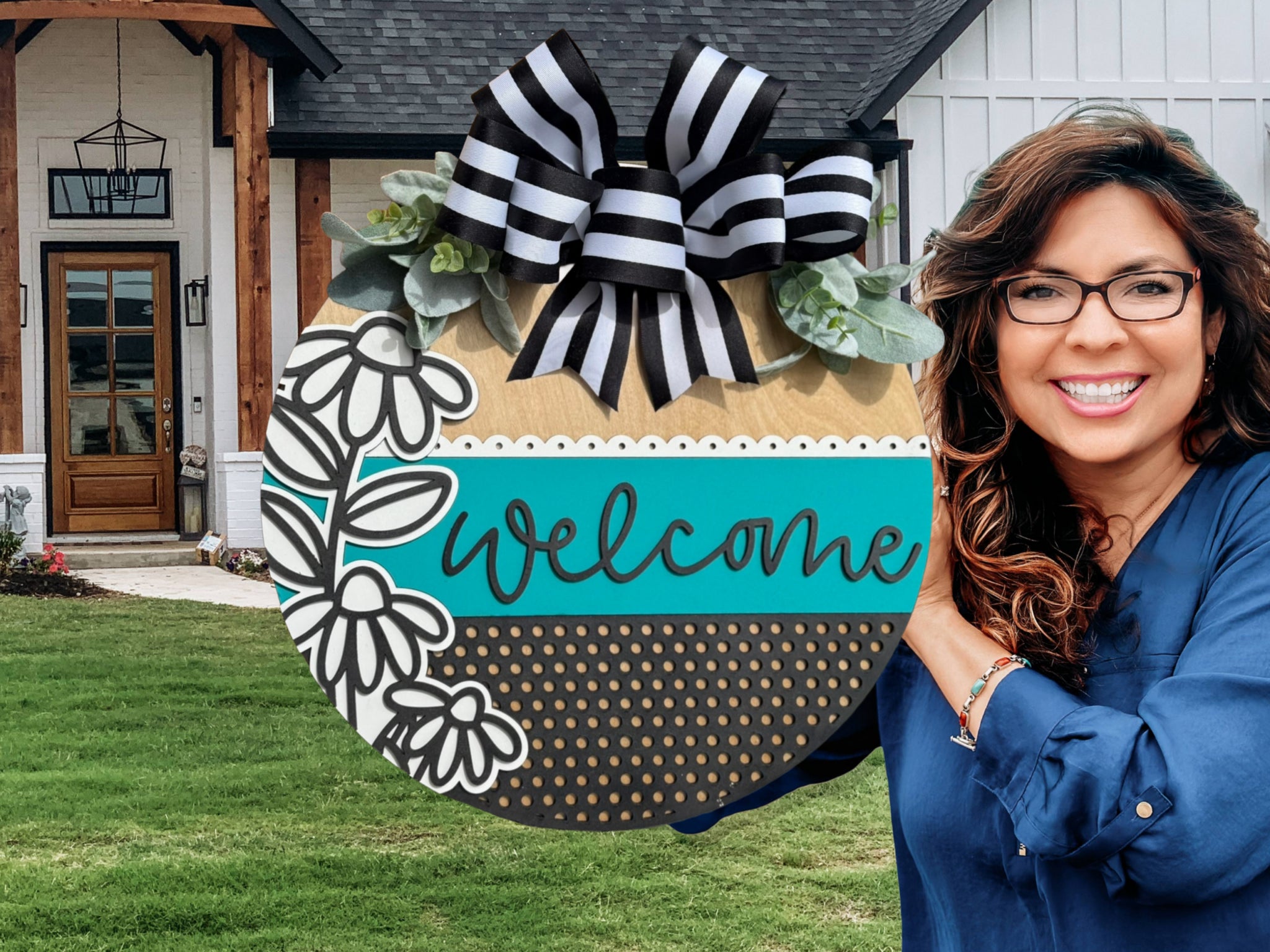 A smiling woman with glasses and long dark hair holds a large round welcome sign decorated with black and white flowers, green leaves, and a striped bow in front of a modern house with wooden and white exterior.