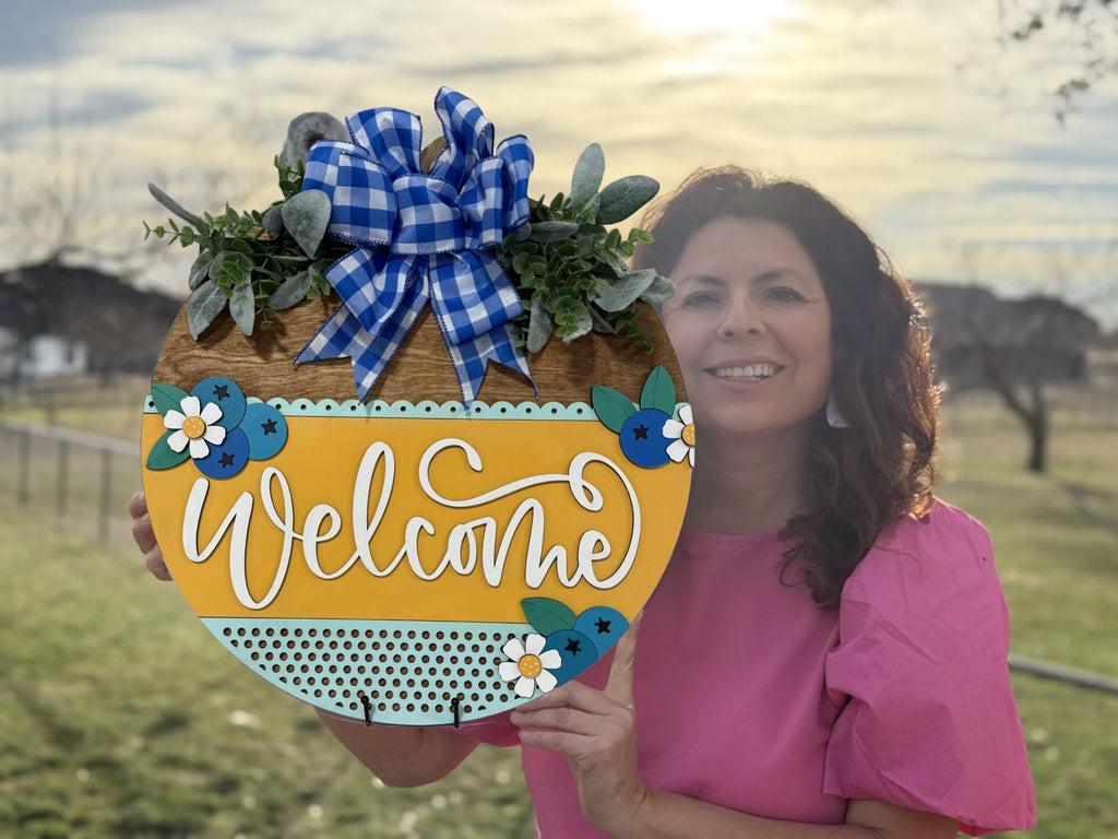 A woman in a pink blouse holds a round wooden welcome sign. The sign features a blue and white checkered bow, greenery, and decorative flowers. Welcome is written in white script on a yellow section. The background shows a sunlit grassy area with trees and houses.
